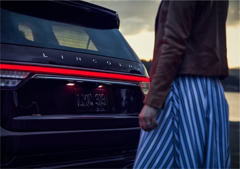 A person is shown near the rear of a 2023 Lincoln Aviator® SUV as the Lincoln Embrace illuminates the rear lights | LaFontaine Lincoln Grand Rapids in Grand Rapids MI