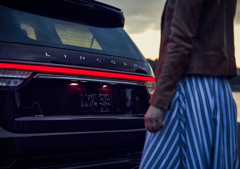 A person is shown near the rear of a 2024 Lincoln Aviator® SUV as the Lincoln Embrace illuminates the rear lights | LaFontaine Lincoln Grand Rapids in Grand Rapids MI